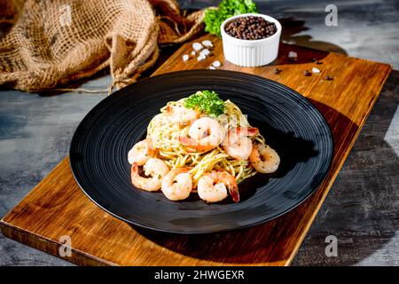 italienische Küche Garnele Aglio Olio Nudel mit schwarzem Pfeffer in einem Gericht auf Holzbrett Seitenansicht auf dunklem Hintergrund Stockfoto