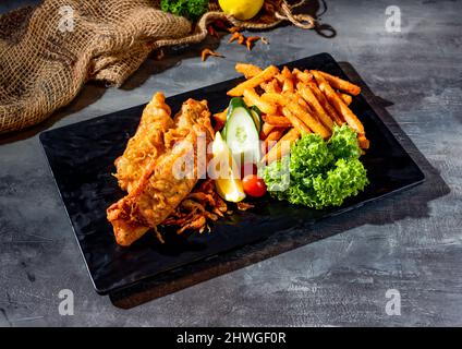 Gebratener roter Tilapia-Fisch mit Salat, Limette und Pommes in einer Beilage auf dunklem Hintergrund Stockfoto