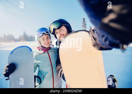 Glückliche Liebhaber Paar Mann und junge Frauen Snowboarder machen Selfie-Foto auf dem Hintergrund des Skigebiets, Sonnenlicht. Stockfoto