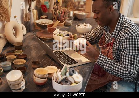 Fröhlicher Keramiker, der am Laptop sitzt und dem Kunden Steingut-Produkte zeigt Stockfoto