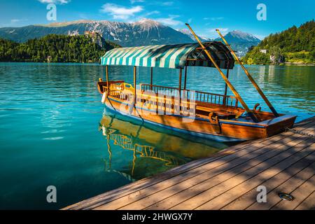 Traditionelles touristisches hölzerne Ruderboot Pletna, das am Pier verankert ist. Holzboot Pletna auf dem berühmten Bleder See und mittelalterliche Burg auf dem Hügel in BA Stockfoto