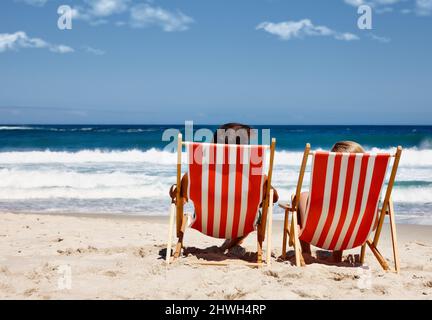 Nicht stören, im Urlaub. Rückansicht eines Paares, das auf den Liegestühlen am Wasserrand liegt. Stockfoto