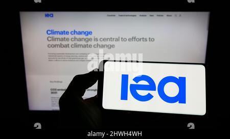 Person mit Mobiltelefon und Logo der Organisation International Energy Agency (IEA) auf dem Bildschirm vor der Webseite. Konzentrieren Sie sich auf die Telefonanzeige. Stockfoto