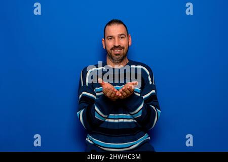 Attraktiver hispanischer Mann mit einem Bart in einem gestreiften Pullover, der etwas mit seinen Handflächen hält und es der Kamera anbietet, während er lächelt Stockfoto