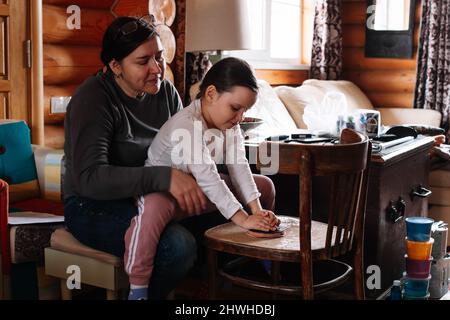 Junges Mädchen, das auf den Knien der Mutter sitzt und der Tochter beibringt, Sand zu schleifen und alte Möbel mit Schleifpapier in der heimischen Werkstatt zu entfernen. Nachhaltig Stockfoto