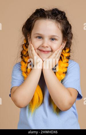 Porträt eines jungen Kindes mit Händen auf den Wangen leicht lächelnd Blick auf die Kamera mit leuchtenden Augen mit gelben Kanekalon Zöpfe auf dem Kopf auf beige Stockfoto