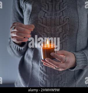 Zünden Sie eine Sojakerze an und halten Sie sie in den Händen. Kerze in einem Glas. Aromatheramia. Duftkerze. Streichelspiel. Kuscheliger grauer Strickpullover. Stockfoto