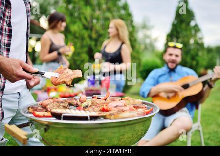 Die Hand des Mannes hält eine Grillzange mit einem saftig leckeren Fleischsteak vor dem Hintergrund eines Grillgrills mit Fleisch und Gemüse und einer Gruppe von Stockfoto