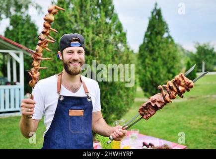 Der junge bärtige Mann in einer Mütze und einer Jeanschürze lächelt und hält einen Spieß mit köstlichem Grillfleisch, das vor dem Hintergrund auf dem Grill zubereitet wird Stockfoto