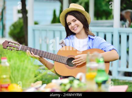 Hübsches Mädchen in einem Strohhut spielt Gitarre und lächelt bei einem Picknick vor dem Hintergrund eines Pavillons Stockfoto