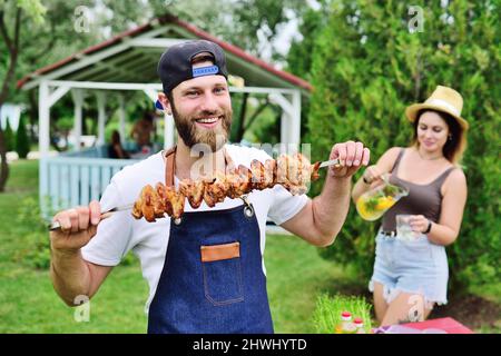 Der junge bärtige Mann in einer Mütze und einer Jeanschürze lächelt und hält einen Spieß mit köstlichem Grillfleisch, das vor dem Hintergrund auf dem Grill zubereitet wird Stockfoto