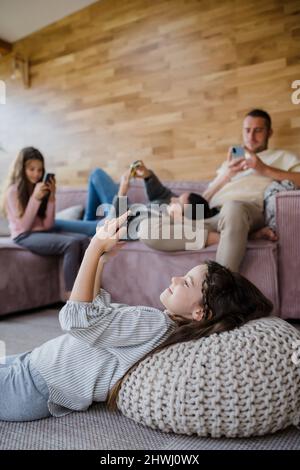 Familie mit zwei kleinen Töchtern, jeder nutzt Handy im Wohnzimmer. Stockfoto