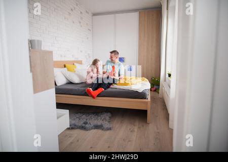 Vater zog seiner kleinen Tochter mit Down-Syndrom verschiedene Socken an, wenn er zu Hause auf dem Bett saß. Stockfoto