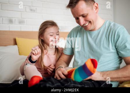 Vater zog seiner kleinen Tochter mit Down-Syndrom verschiedene Socken an, wenn er zu Hause auf dem Bett saß. Stockfoto