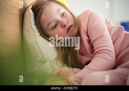 Trauriges kleines Mädchen mit Down-Syndrom, das zu Hause auf dem Bett liegt. Stockfoto