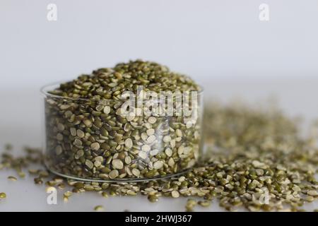 Teilen Sie die Linsen aus monognen Bohnen in einer kleinen Glasschüssel. Auf weißem Hintergrund geschossen, mit Haufen gespaltener Moong-Bohnen sind verstreut. Stockfoto