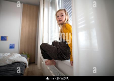 Trauriges kleines Mädchen mit Down-Syndrom, das zu Hause am Fenster sitzt. Stockfoto