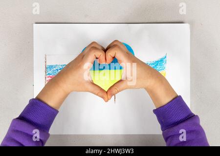 Ein Kind macht mit den Händen ein Herzzeichen über einer Zeichnung von zwei Flaggen der Ukraine und Russlands aus der Nähe. Blick von oben. Kinder gegen Krieg. Stockfoto