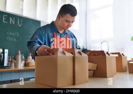 Fröhlicher junger Kellner mit Down-Syndrom, der im Take-Away-Restaurant arbeitet, Konzept der sozialen Eingliederung. Stockfoto