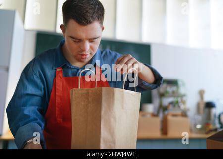 Fröhlicher junger Kellner mit Down-Syndrom, der im Take-Away-Restaurant arbeitet, Konzept der sozialen Eingliederung. Stockfoto