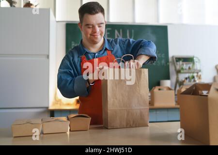 Fröhlicher junger Kellner mit Down-Syndrom, der im Take-Away-Restaurant arbeitet, Konzept der sozialen Eingliederung. Stockfoto