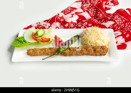 Türkischer adana Kebab mit Reis und Salat in einem Gericht isoliert auf bunten Tischtuch Seitenansicht auf grauem Hintergrund Stockfoto