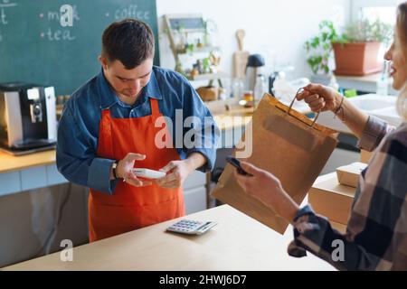 Fröhlicher junger Down-Syndrom-Kellner, der kontaktlose Smartphone-Zahlung vom Kunden im Take-Away-Restaurant nimmt. Stockfoto