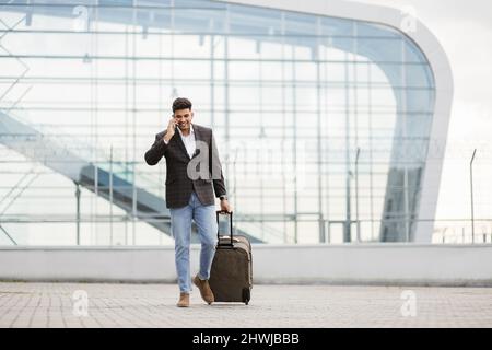 Angenehmer, junger arabischer Geschäftsreisender, der am Telefon telefoniert, während er mit Koffern aus dem modernen Flughafen läuft Stockfoto