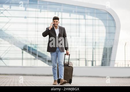 Angenehmer, junger arabischer Geschäftsreisender, der am Telefon telefoniert, während er mit Koffern aus dem modernen Flughafen läuft Stockfoto