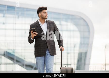 Junger gutaussehender männlicher indischer Geschäftsmann in formeller Jacke, der in der Nähe eines modernen Gebäudes steht und draußen auf dem Smartphone schreibt. Arabischer Mann, der draußen telefoniert Stockfoto
