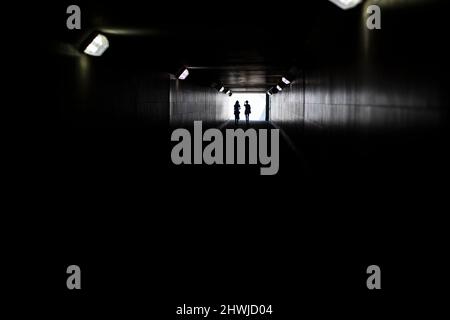 Zwei Silhouetten am Ende des Tunnels. Licht in der Ferne. Figuren von kleinen Frauen in kontrastierendem Licht. Lampen in langer Unterführung. Stockfoto