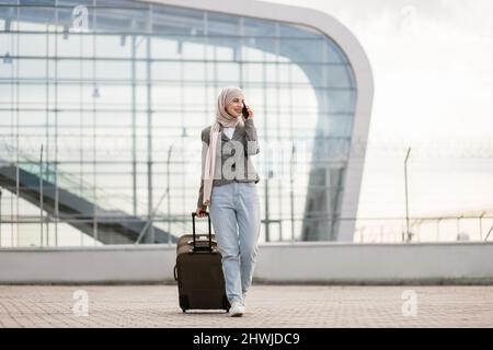 Porträt einer glücklichen muslimischen arabischen Frau, die einen Hijab und legere Kleidung trägt, im Freien mit einem Koffer und einem Telefongespräch unterwegs ist und mit dem Flugzeug auf dem modernen Flughafen unterwegs ist. Stockfoto