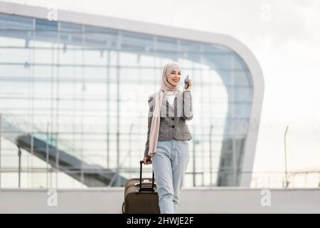 Flugreise und Geschäftskonzept. Hübsche Reisende Touristen-arabische Frau in Hijab, die Koffer und Telefon trug und in der Nähe des modernen Flughafenterminals ging Stockfoto