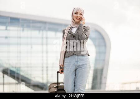 Flugreise und Geschäftskonzept. Hübsche Reisende Touristen-arabische Frau in Hijab, die Koffer und Telefon trug und in der Nähe des modernen Flughafenterminals ging Stockfoto