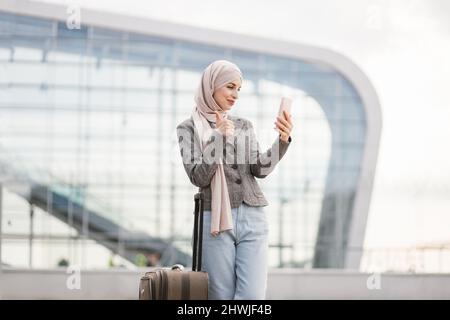 Junge hübsche muslimische Frau, die ihr Mobiltelefon hält, während sie mit einem Lächeln zu ihrem Freund anruft. Arabische Geschäftsfrau, die vor einem modernen Gebäude mit Mobiltelefon und Videokonferenzanruf steht Stockfoto