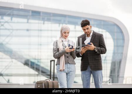 Porträt eines glücklichen muslimischen Paares, einer Frau in Hijab und manin-Jacke, die mit Pässen und Tickets im Freien unterwegs ist, einen Koffer trägt und sich gegenseitig unterredet, mit dem Flugzeug auf dem modernen Flughafen unterwegs ist. Stockfoto