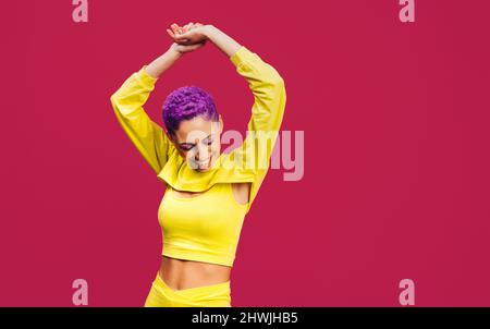 Frau mit lila Haaren tanzt fröhlich im Studio. Glückliche junge Frau lächelt und hat Spaß vor einem rosa Hintergrund. Modische junge Frau s Stockfoto