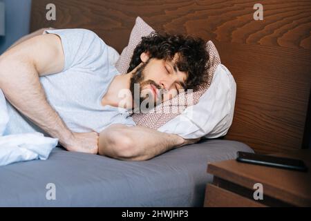 Nahaufnahme des müden bärtigen jungen Mannes, der friedlich auf der Seite in einem großen komfortablen Bett unter einer weißen Decke schläft, Stockfoto