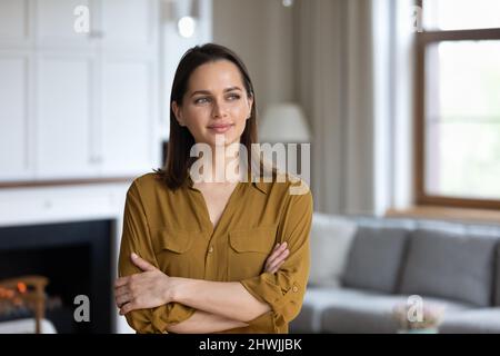Nachdenkliche Frau, die im Wohnzimmer steht, lächelt und blickt in die Ferne Stockfoto