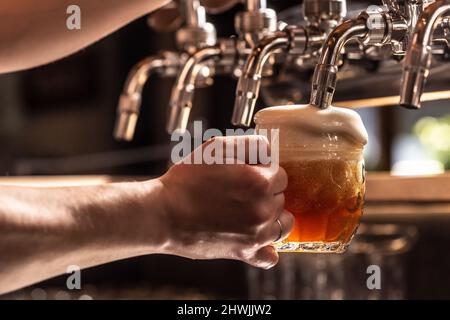 Frau hält einen Becher Bier, der aus dem Hahn mit Schaum gefüllt ist, der aus dem Glas schwebt. Stockfoto