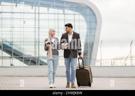 Porträt eines glücklichen muslimischen Paares, einer Frau in Hijab und manin-Jacke, die mit Pässen und Tickets im Freien unterwegs ist, einen Koffer trägt und sich gegenseitig unterredet, mit dem Flugzeug auf dem modernen Flughafen unterwegs ist. Stockfoto