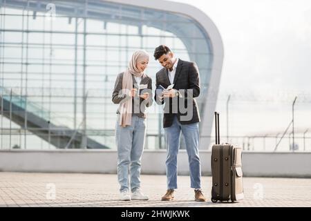 Business-, Reise- und Mitarbeiterkonzept. Glückliches Geschäftsehepaar, muslimische Frau im Hijab und arabischer Mann, der Koffer vor dem Flughafen trägt, Pässe und Tickets hält. Frau, die telefoniert Stockfoto