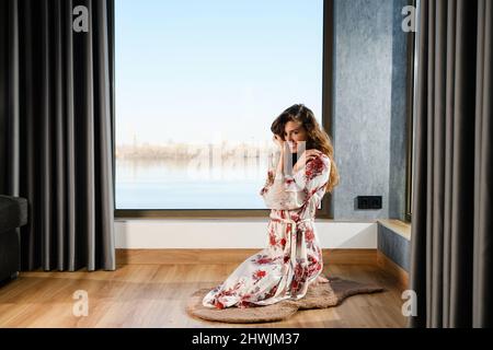 Frau in japanischem Seidenkleid sitzt auf Holzboden der modernen Wohneinrichtung mit Panoramafenster, Morgenübungen Stockfoto