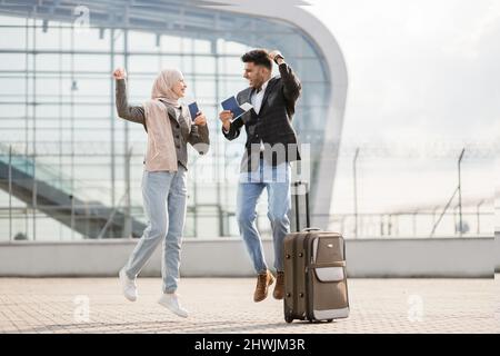 Ein glückliches, aufgeregtes Paar, eine muslimische Dame im Hijab und ein hübscher arabischer Mann, sind froh und springen zusammen, stehen vor dem modernen Flughafenterminal und halten Pässe und Tickets in der Hand. Reisekonzept für Freunde Stockfoto