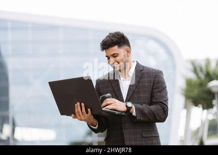 Fokussierter junger erfolgreicher arabischer Geschäftsmann in formeller Kleidung, der vor einem modernen Gebäude steht und auf einem Laptop im Internet surft. Selbstbewusster bärtiger Mann, der beim Arbeiten im Freien frische Luft genießt. Stockfoto