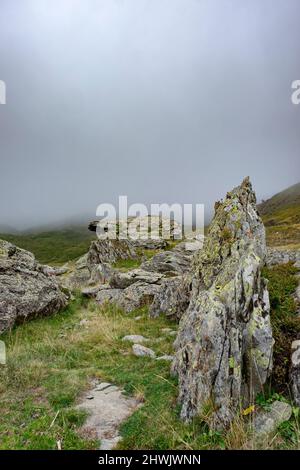 Hochgebirgslandschaft mit granitischen Felsen, die sich im Vordergrund mit Flechten bedecken, und einer dicken Nebelbank, die die Berge im Hintergrund bedeckt, verti Stockfoto