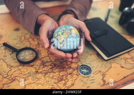 Globus, die ganze Welt in Händen und Kompass, Lupe und Buch auf der Routenkarte auf dem Tisch. Reise-, Abenteuer- und Entdeckungskonzept. Stockfoto