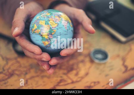 Globus, die ganze Welt in Händen und Kompass, Lupe und Buch auf der Routenkarte auf dem Tisch. Reise-, Abenteuer- und Entdeckungskonzept. Stockfoto
