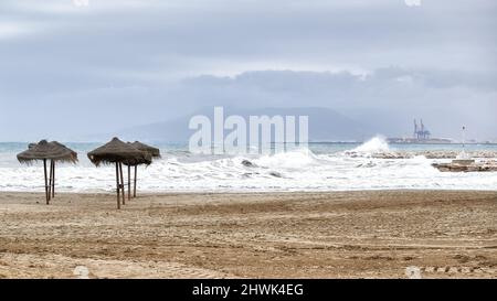 Schönes Bild des Strandes in der Stadt Malaga, Spanien im Winter mit leeren Sonnenschirmen, starken Wellen, die gegen das Wellenbrecher und den por krachen Stockfoto