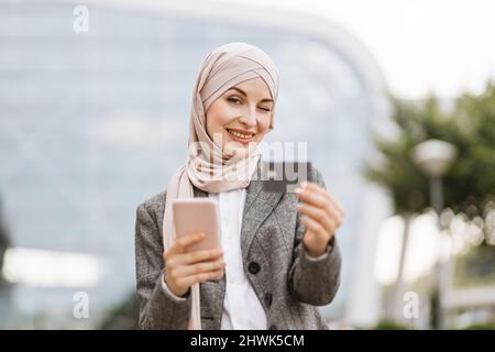 Ziemlich glückliche muslimische Dame im Hijab lächelt an der Kamera, steht draußen mit Kaffee zu gehen und in den Händen telefonieren. Sympathische arabische Frau im Kopftuch, die mit Kaffee zum Mitnehmen und Telefon auf der Straße unterwegs ist Stockfoto
